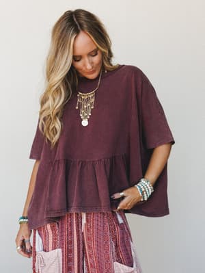 A relaxed, oversized ruffle-hem top in washed mulberry styled over a printed boho skirt, layered with gold coin jewelry and stacked beaded bracelets — the kind of effortless outfit that looks like it came together naturally but definitely didn't.