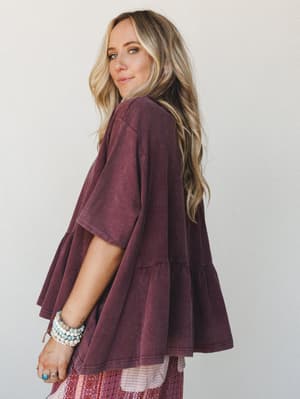 A blonde model shot from a three-quarter back angle, glancing over her shoulder in a relaxed acid-wash mulberry tiered ruffle top — styled with a printed boho skirt and stacked beaded bracelets, the whole look feels like a cozy Saturday with somewher