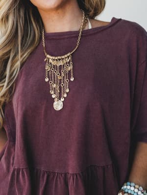 A close-up of a relaxed mulberry ruffle-hem top styled with a statement gold fringe coin necklace — the kind of effortless pairing that looks like you thought about it for exactly the right amount of time.