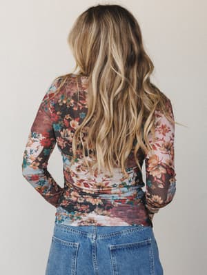 Shot from behind, a model in a long-sleeve floral mesh top tucked into medium-wash jeans — the kind of top that does all the talking while you just show up.