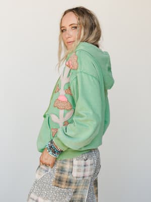 A mint green hoodie with large floral appliqué and sequin embellishments down the front and sleeve, styled with mixed plaid cargo pants and stacked beaded bracelets — shot from a three-quarter back angle with a soft, lived-in ease.