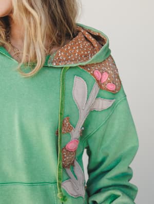 A close-up of a washed green hoodie with artisan-style fabric appliqué — botanical leaf and berry motifs in sage, brown, and dusty pink layered across the shoulder and chest, giving it that one-of-a-kind, made-with-intention feel.