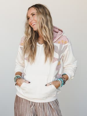 A cream hoodie with soft floral patchwork appliqué in blush, taupe, and gold styled with striped wide-leg pants and stacked turquoise bracelets — the kind of cozy-but-collected outfit that makes a Saturday feel like a whole mood.