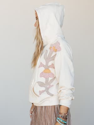 A side-profile shot of a model in a cream hoodie with large-scale floral appliqué detail — soft, handmade-feeling, and the kind of thing you'd wear to the farmer's market and still feel put-together in.