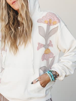 A cream hoodie with oversized floral appliqué in dusty pink and taupe takes center stage here — the kind of piece that looks just as good with yesterday's jeans as it does with something intentional, styled with a stack of turquoise and beaded bracel