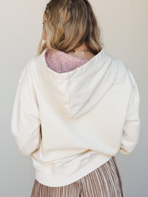 A back-view shot of a cream hoodie styled over a pink printed top and a pleated bronze skirt — the kind of effortless layering that looks accidental but really isn't.