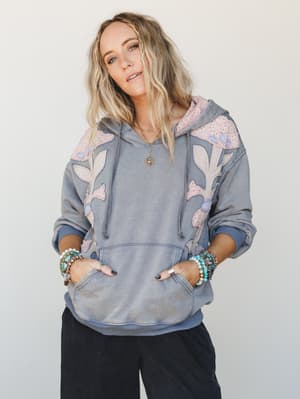 A washed denim-blue hoodie with soft floral appliqué detailing worn casually with black wide-leg pants — the kind of piece that makes a lazy Saturday feel intentional.