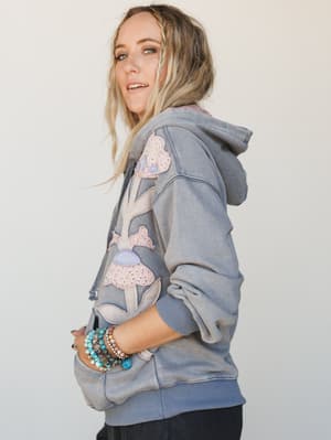 A side-profile shot of a model wearing an oversized washed denim hoodie covered in soft floral and mushroom appliqué patches, styled with a stacked mix of turquoise and beaded bracelets — the kind of sweatshirt that feels like a hug and a personality
