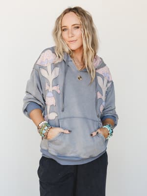 A washed denim-blue oversized hoodie with large floral appliqué patches in blush and lavender worn hands-in-pocket with stacked boho bracelets and a gold pendant necklace — the kind of sweatshirt that makes getting dressed feel effortless and a littl
