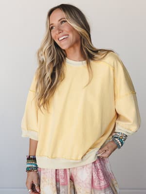 A sun-warmed banana yellow pullover sweatshirt styled with a floral skirt and stacked beaded bracelets — the kind of outfit that looks effortless at 10am and still good at 6pm.