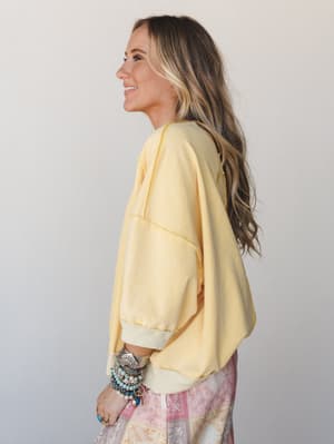 A side-profile shot of a model in a soft butter-yellow cropped sweatshirt with visible seam detailing, layered over a pink paisley skirt and stacked with boho bracelets — the kind of effortless, sunny outfit you'd actually reach for on a good day.