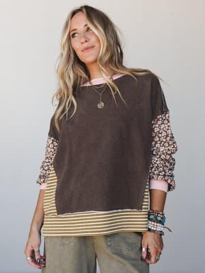 A relaxed mixed-media top in coffee brown with floral sleeves, layered over a striped hem layer and pink ribbed collar — styled with olive wide-leg pants and a stack of boho bracelets for that perfectly collected, not-too-trying look.