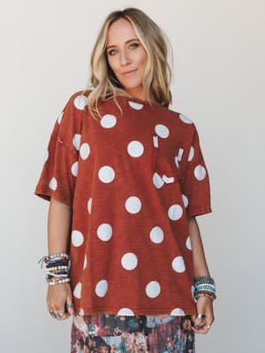An oversized rust-toned tee with bold white polka dots gets layered over a floral midi skirt, styled with stacks of beaded bracelets — the kind of effortless, personality-forward combo that looks like it took zero effort and all the intention.