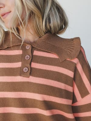 A close-up detail shot of a striped knit polo sweater in warm brown and dusty pink, showing a ribbed collar, button placket, and cozy knit texture — the kind of piece that makes a Tuesday feel a little more intentional.