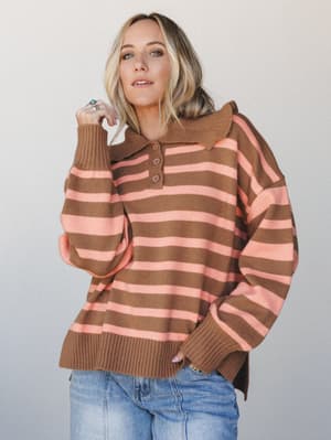 An oversized striped polo sweater in warm camel and coral pink worn with light wash jeans — the kind of cozy, personality-forward piece that feels equally at home on a coffee run or a casual Friday.