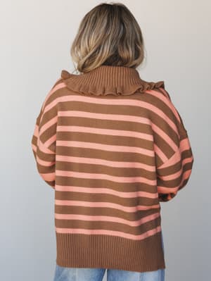 Shot from the back, a model wears an oversized striped knit sweater in warm camel and coral pink, with a standout ruffled turtleneck collar — the kind of cozy piece that feels as good as it looks.