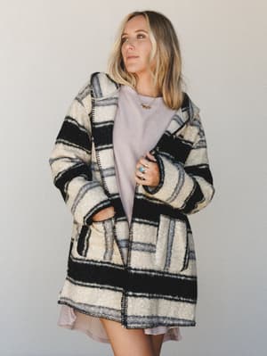 A relaxed, dreamy studio shot of a model layering an oversized striped wool-blend coat over a soft lavender mini dress — the kind of outfit that looks effortless but clearly wasn't an accident.
