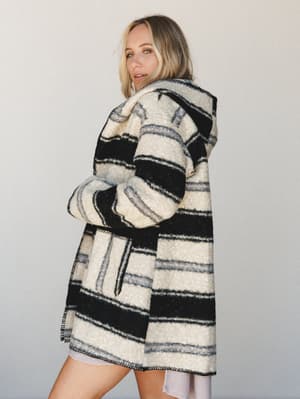A blonde model looks back over her shoulder wearing an oversized hooded blanket coat in cream and black stripe — the kind of jacket that makes you want to take a road trip with nowhere to be.
