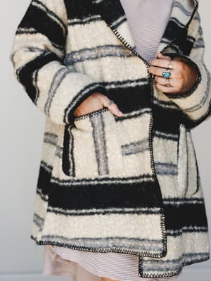 A close-up detail shot of a chunky striped wool-blend jacket layered over a blush knit dress, styled with stacked silver and turquoise rings — the kind of jacket that makes you want to take a long walk somewhere with good coffee.