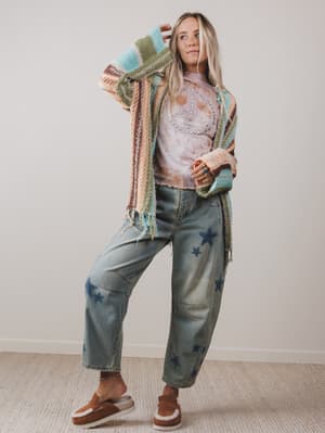 Person wearing a colorful cardigan, patterned shirt, starry jeans, and brown sandals against a plain background.