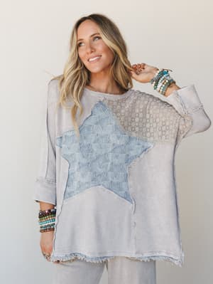 A relaxed oversized top in washed light ash with a large star appliqué and mixed-fabric patchwork panels — worn with stacks of beaded bracelets and an easy smile, it's the kind of top that looks like it has a story.