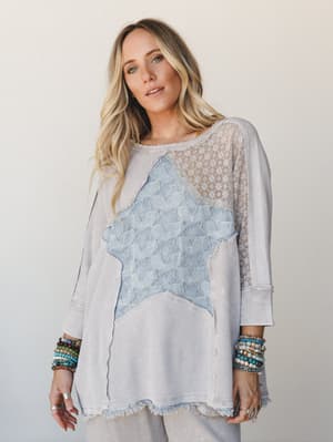 A relaxed, mixed-media top in washed light ash layered with a star-print denim patch appliqué and sheer lace panel — styled with stacks of beaded bracelets for that effortless, lived-in boho feel.