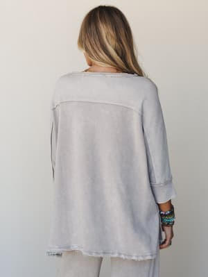 A back-view shot of a relaxed, oversized top in washed light ash — the mixed fabric panels, raw hem, and stack of beaded bracelets give it that effortlessly lived-in, boho-casual feel.