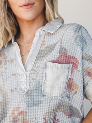 A close-up of a washed grey waffle-knit top with a faded floral print, collared v-neckline, and chest pocket — the kind of top that looks like it's been loved for years and somehow only gets better.