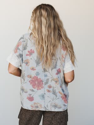 A relaxed, short-sleeve waffle-knit top in dusty blue-grey with a faded botanical floral print, styled from the back and paired with dark leopard cargo pants — effortlessly lived-in and a little unexpected.
