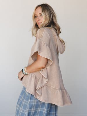 Shot from the back at a three-quarter angle, this model shows off a washed almond hoodie tee with tiered ruffle hem paired with a blue plaid skirt — the kind of effortless, thrown-together outfit that somehow looks completely intentional.