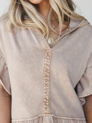 A close-up detail shot of a washed almond hoodie tee with a decorative embroidered front placket and ruffle hem, styled with layered gold and stone necklaces for an effortless, lived-in feel.