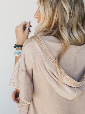 A close-up detail shot of a washed almond hoodie tee, shot from behind to highlight the braided trim along the hood and the flutter sleeve — styled with a stack of turquoise and beaded bracelets that feel collected, not coordinated.