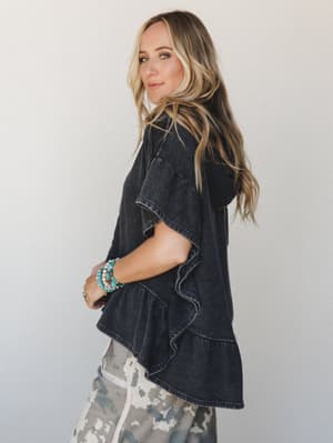 A washed-black hooded tee with ruffle hem details is styled with camo joggers and a stack of turquoise bracelets — the kind of effortless mix that looks intentional without trying too hard.