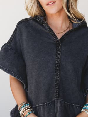 A close-up of a washed charcoal hoodie top with crochet trim detailing down the center placket, styled with layered beaded bracelets and a gold chain necklace — the kind of piece that looks equally at home on a coffee run or a lazy weekend road trip.