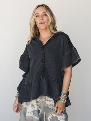 A relaxed, washed-black hooded top with tiered ruffle hem and crochet trim detail, styled with camo wide-leg pants and stacked turquoise bracelets — effortless boho energy that feels lived-in and totally intentional.
