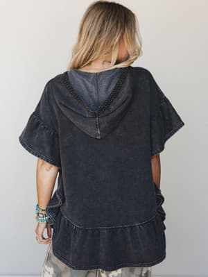 A back-view shot of a washed black hooded tee with ruffle-tiered hem and flutter sleeves, styled with camo pants and stacked turquoise bracelets against a clean studio backdrop — casual and boho in equal measure.