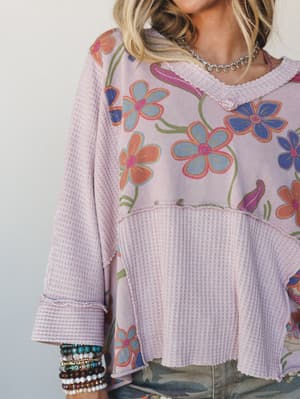 A close-up of a mixed-media floral top in soft mauve, combining a retro large-scale floral print panel with waffle-knit fabric blocking — styled with a stack of beaded bracelets and a silver chain necklace for that effortlessly collected, personal-st