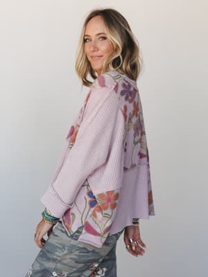 A side-back view of a mixed-media waffle knit top in soft lavender-mauve with bold retro floral print panels, paired with camo jeans and layered jewelry — the kind of top that makes a Tuesday feel intentional.