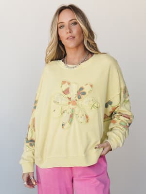 A soft butter-yellow oversized sweatshirt with a patchwork floral appliqué on the chest and printed floral sleeves, styled with bright pink pants for a playful, lived-in look.