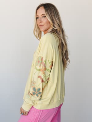 A soft butter-yellow pullover sweatshirt with retro floral graphic sleeves styled with pink jeans — the kind of top that looks equally at home on a coffee run or a slow Saturday morning.