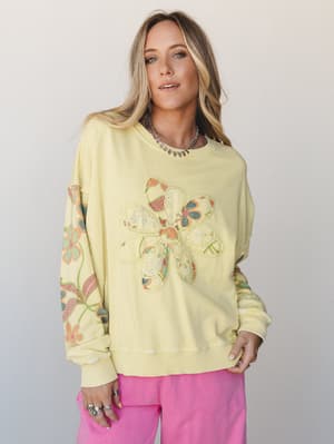A soft lime yellow oversized sweatshirt with a patchwork floral appliqué at the center and embroidered floral sleeves, styled with bright pink pants for a playful, lived-in boho vibe.