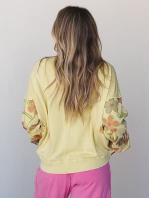 A back-facing shot of a soft lime yellow pullover top with voluminous floral-printed sleeves in retro orange, green, and pink tones, paired with bright pink wide-leg pants against a clean light grey studio backdrop.