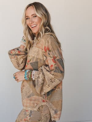 A smiling model in a patchwork floral top with wide bell sleeves, layered with beaded bracelets and rings — the kind of outfit that looks like it came together effortlessly but clearly didn't.