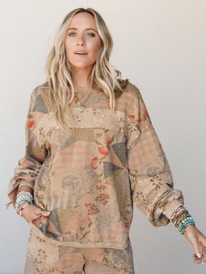 A patchwork floral pullover top in warm latte tones layered with eyelet trim panels — worn with stacked beaded bracelets and a relaxed ease that feels like your favorite worn-in piece finally got a glow-up.