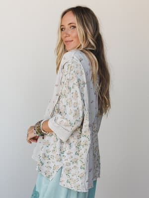 A model glances back over her shoulder wearing a relaxed floral top in stone grey with soft cream blooms, layered with stacked bracelets and mint wide-leg pants — the kind of outfit that looks effortless but absolutely wasn't an accident.