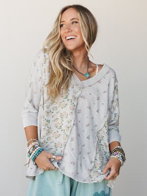 A smiling blonde model in a relaxed floral patchwork top with raw-edge seaming, layered boho bracelets, and a turquoise butterfly necklace — the kind of outfit that looks like it just happened and somehow looks perfect.