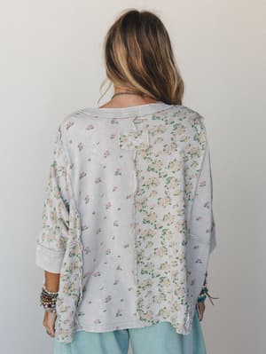 A back-view shot of a relaxed floral sweatshirt top in washed stone grey, featuring mixed floral panels and exposed seam detailing — the kind of piece that looks like it was meant to be lived in.