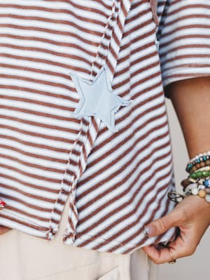 A close-up detail shot of a rust-striped knit top with a frayed light blue star appliqué at the hem, styled with cream bottoms and a stack of mixed bead bracelets — casual and a little nostalgic in the best way.