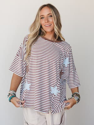 A relaxed, oversized striped top with pale blue star appliqués scattered across the front, styled with stacked beaded bracelets and neutral pants — the kind of effortless, personality-forward piece that does all the talking.
