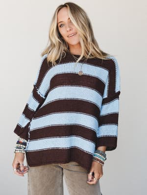 An oversized striped knit sweater in mocha and powder blue worn with relaxed corduroy pants — stacked bracelets and layered necklaces give it that effortless, collected feel.